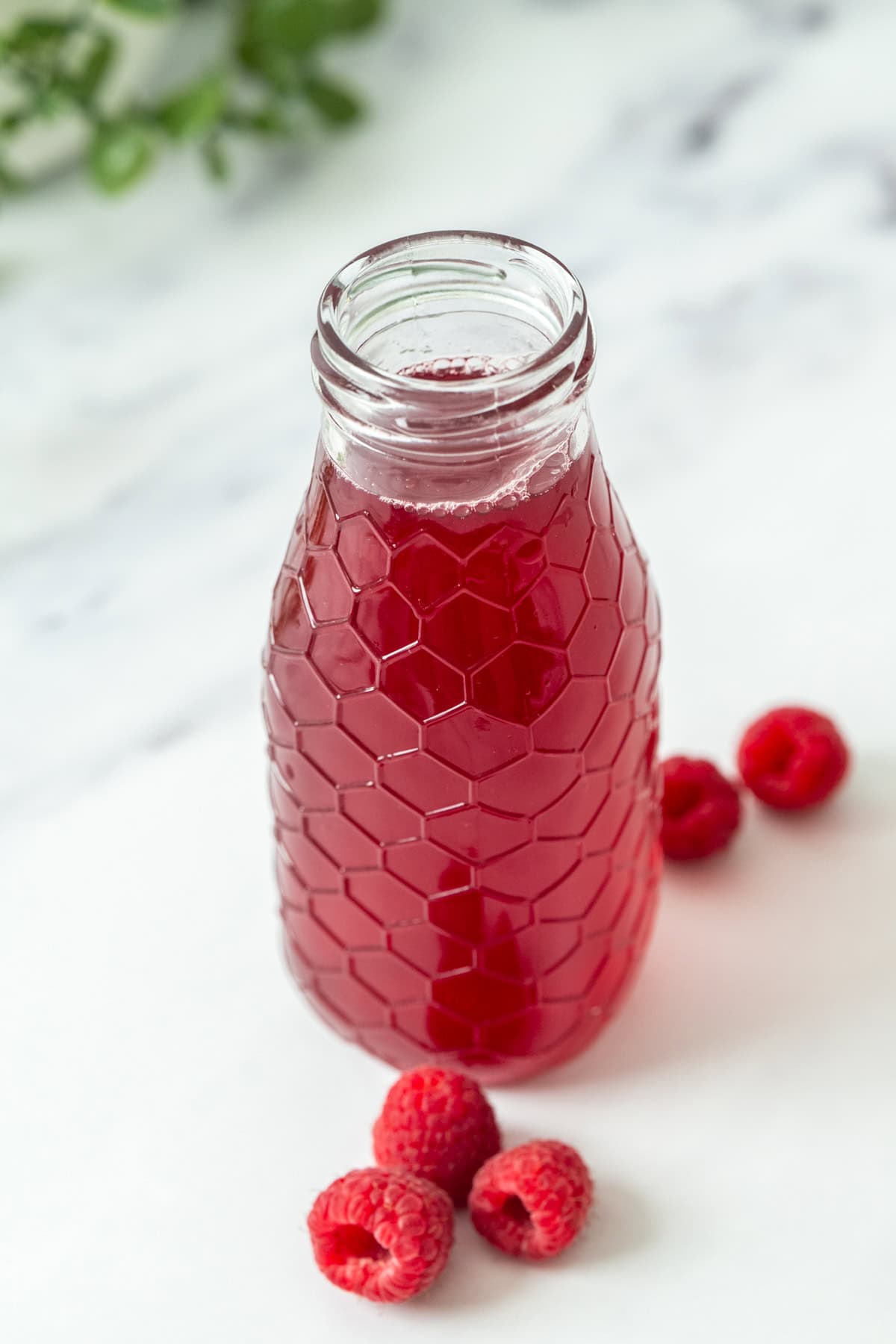 A clear glass bottle filled with raspberry syrup, featuring a honeycomb pattern, sits on a white surface next to a few fresh raspberries. The background is softly blurred.