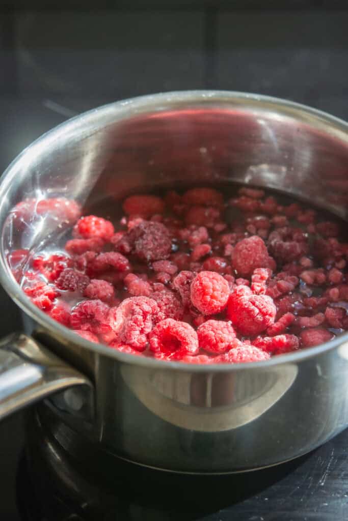 A stainless steel saucepan filled with frozen raspberries is heating on a stovetop, with sunlight highlighting the bright red berries.