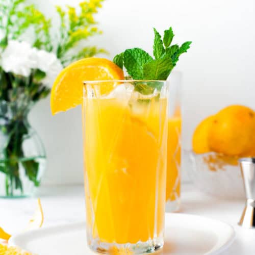 A tall glass of orange juice with ice, garnished with a lemon wedge and mint sprig, sits on a white plate. In the background are fresh flowers, whole lemons, and a glass bowl.