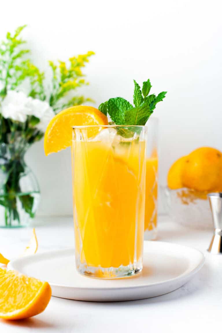 A tall glass of orange juice with ice, garnished with a lemon wedge and mint sprig, sits on a white plate. In the background are fresh flowers, whole lemons, and a glass bowl.