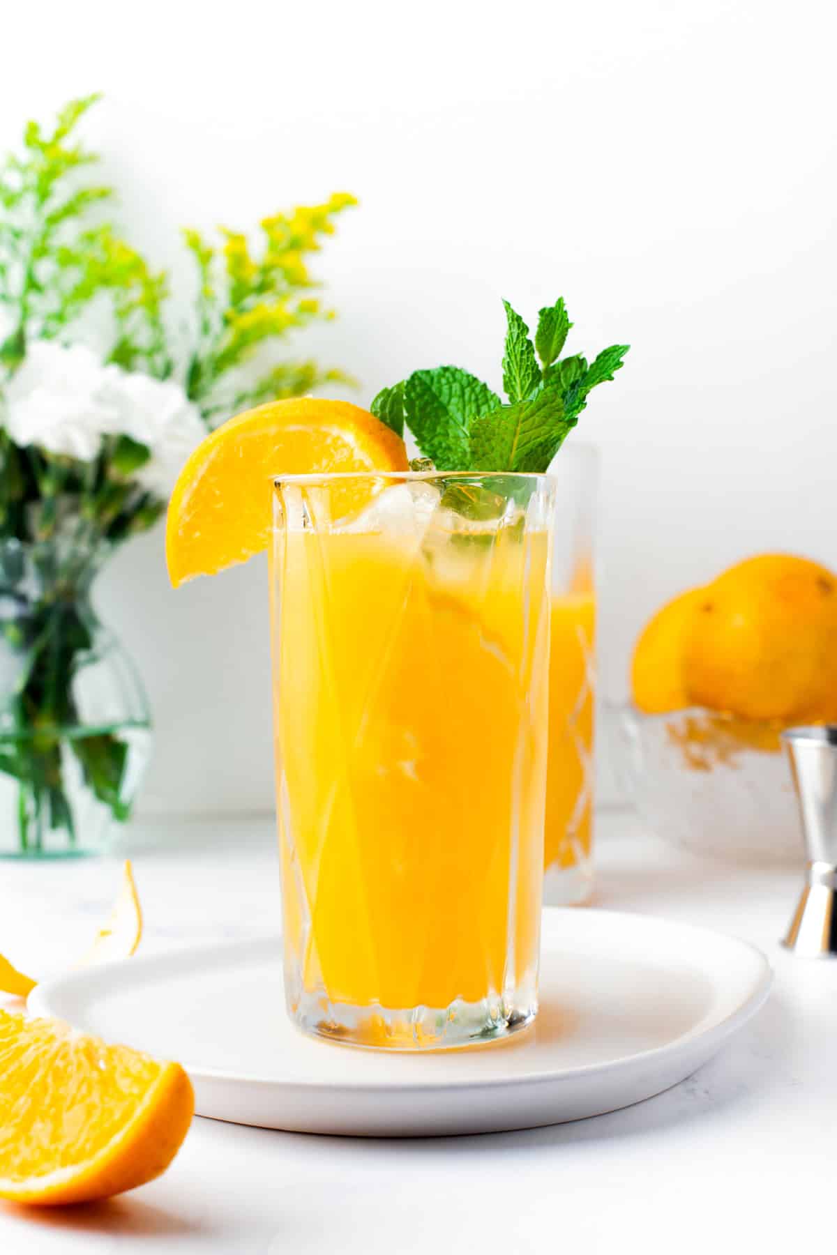 A tall glass of orange juice with ice, garnished with a lemon wedge and mint sprig, sits on a white plate. In the background are fresh flowers, whole lemons, and a glass bowl.