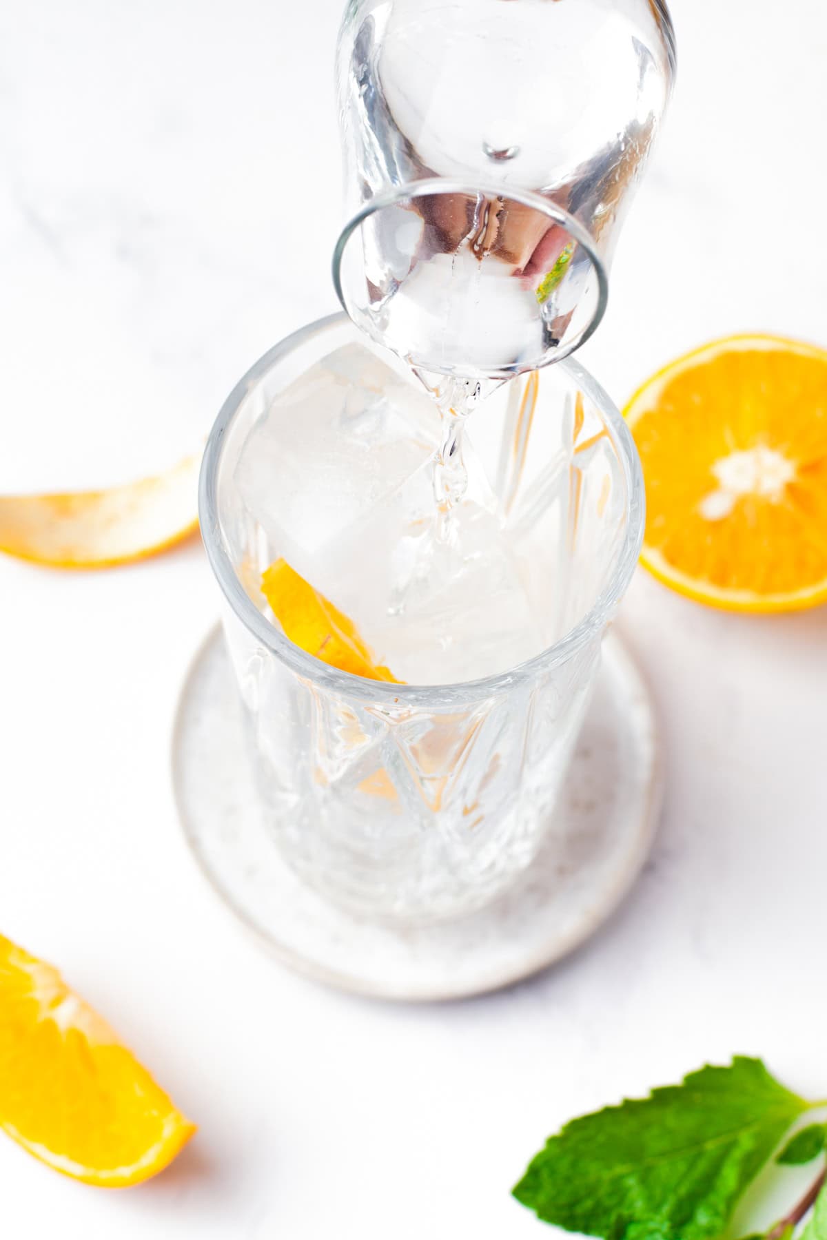 A glass with ice and an orange slice is being filled with clear liquid from a small jug. Orange slices and mint leaves are placed nearby on a white surface.