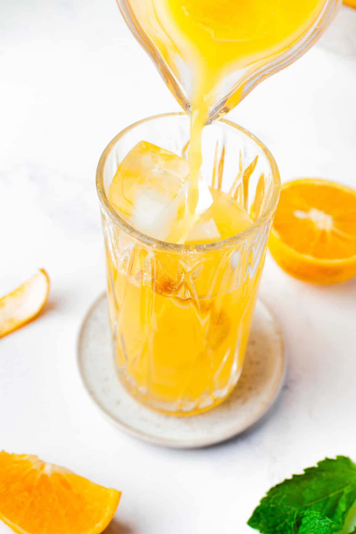 Orange juice being poured into a glass filled with ice cubes, surrounded by fresh orange slices and a green mint leaf on a white surface.