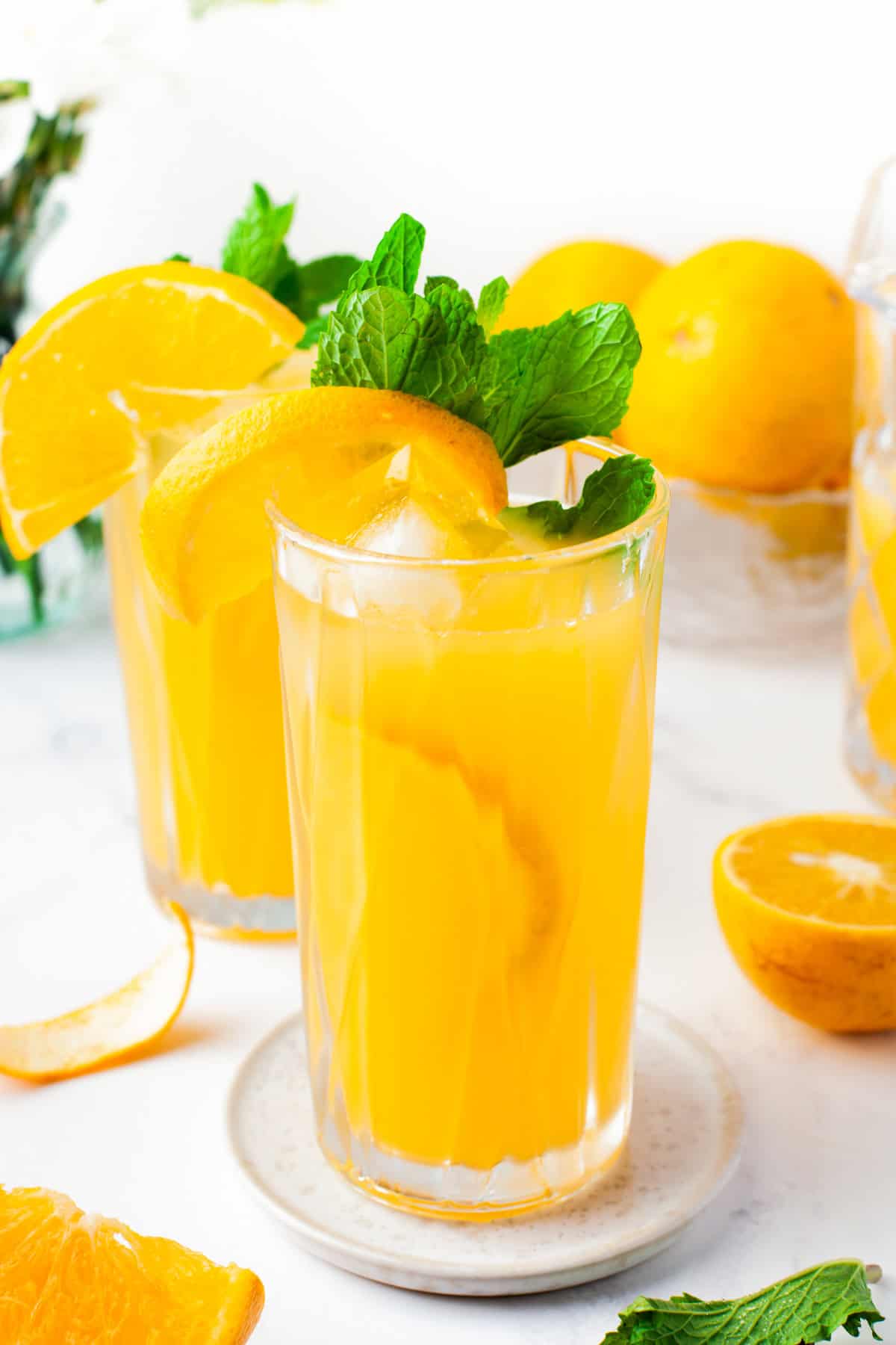 Two tall glasses of orange drink with ice, garnished with orange slices and fresh mint leaves, sit on a white plate. Whole and halved oranges are in the background on a bright, white surface.