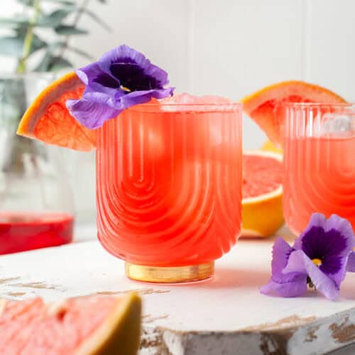 A vibrant pink grapefruit cocktail in a textured glass, garnished with a purple flower and a grapefruit wedge. Another similar drink and grapefruit pieces are in the background, with a blurred leafy plant.