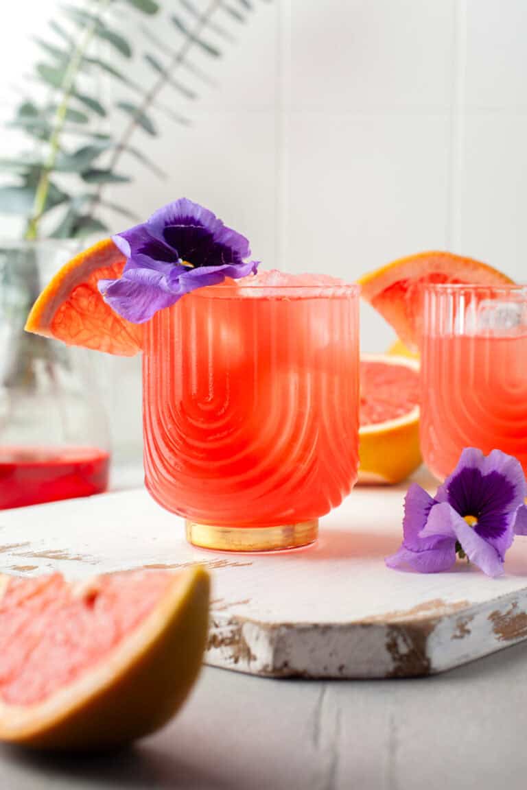 A vibrant pink grapefruit cocktail in a textured glass, garnished with a purple flower and a grapefruit wedge. Another similar drink and grapefruit pieces are in the background, with a blurred leafy plant.