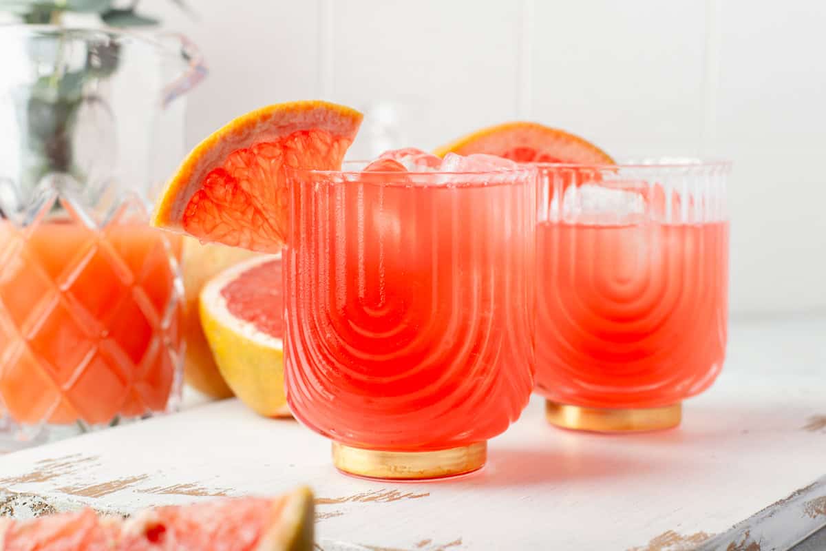 Two glasses of bright pink grapefruit juice with ice and grapefruit wedges as garnish, sitting on a white wooden surface. A jug of juice and a halved grapefruit are in the background.