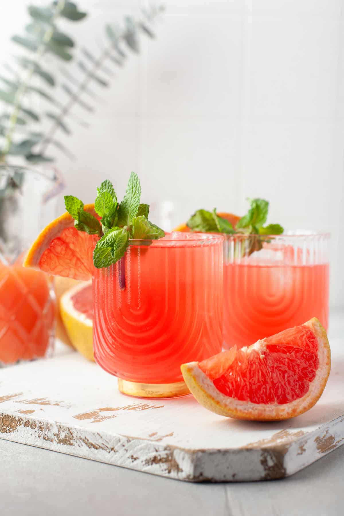 Two glasses of pink grapefruit cocktail garnished with fresh mint and grapefruit slices, placed on a white wooden board with a grapefruit wedge in the foreground. A glass pitcher and greenery are blurred in the background.