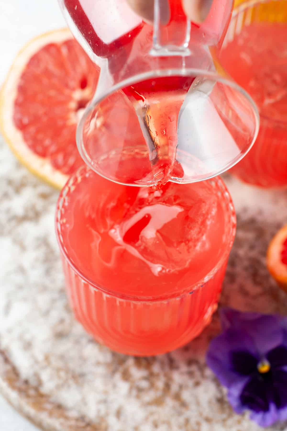 A close-up of a glass pitcher pouring bright pink grapefruit juice into a glass with ice. Sliced grapefruit and a purple flower are on a textured surface beside the drink.