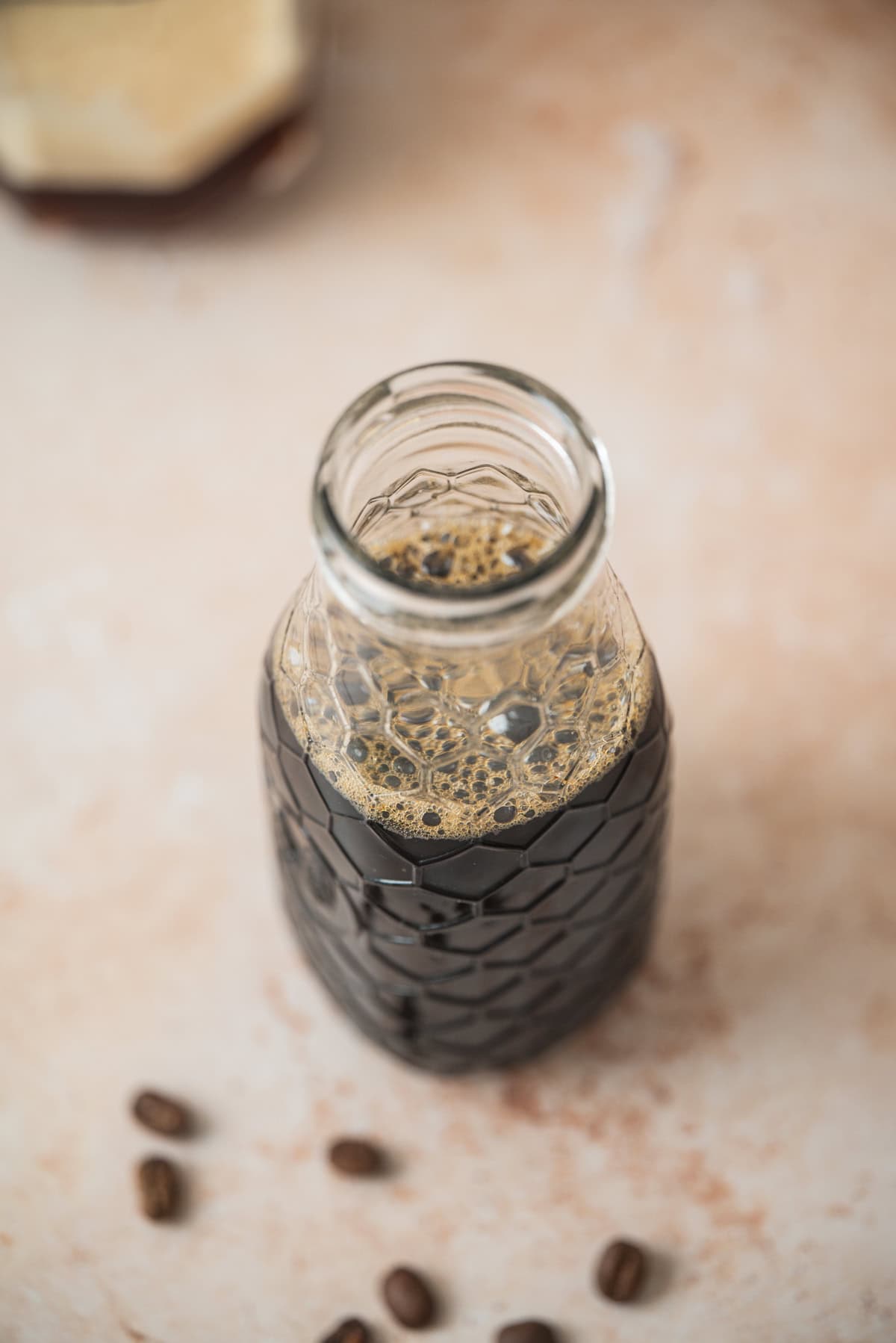 A glass bottle filled with Homemade Virgin Coffee Liqueur sits on a light, textured surface, with a few coffee beans scattered nearby. Bubbles dot the dark liqueur’s surface, hinting at its rich aroma and freshly crafted essence.