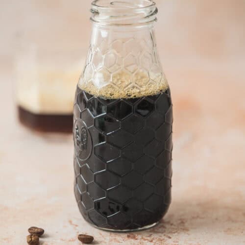 A clear glass bottle with a honeycomb pattern is filled with dark Homemade Virgin Coffee Liqueur. Several coffee beans are scattered in front, and a blurred glass with foam is in the background.