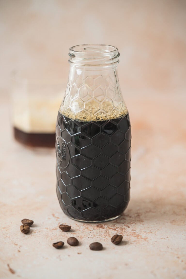 A clear glass bottle with a honeycomb pattern is filled with dark Homemade Virgin Coffee Liqueur. Several coffee beans are scattered in front, and a blurred glass with foam is in the background.