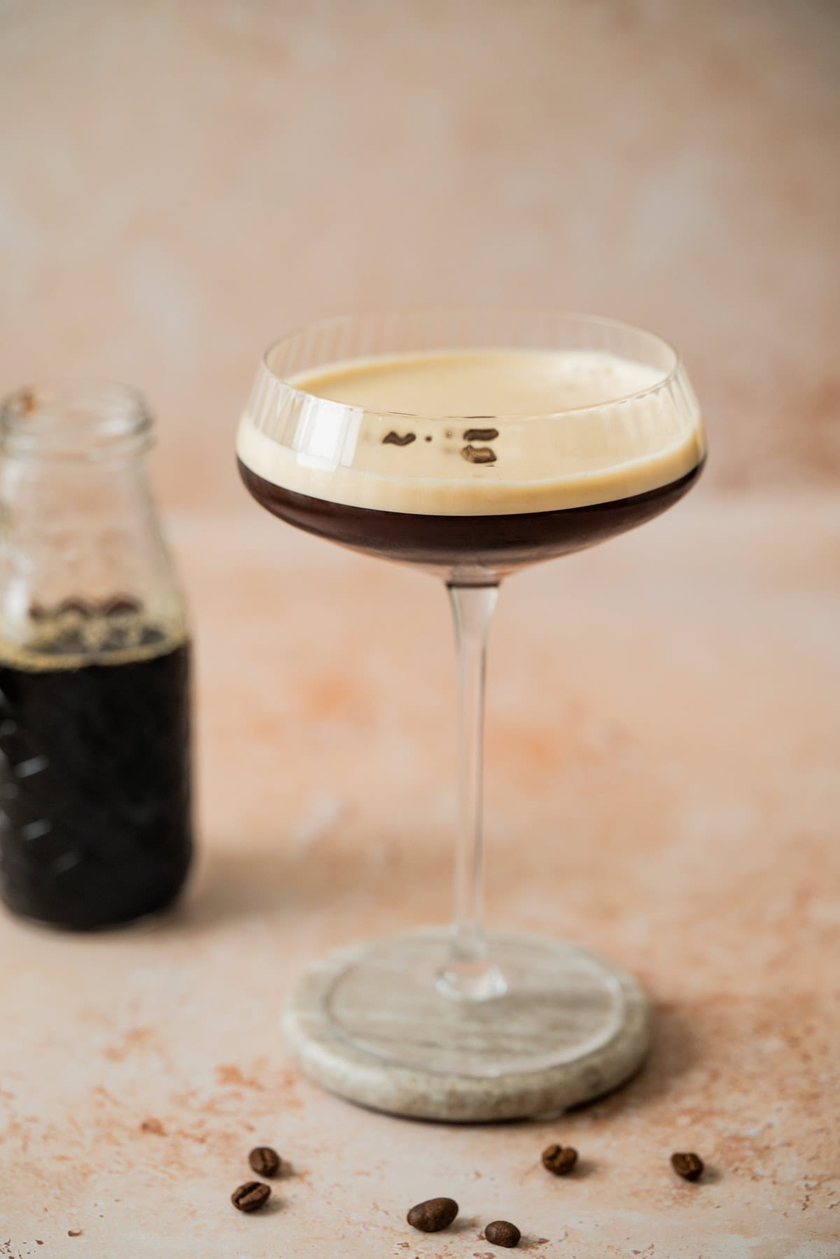 A coupe glass filled with a dark Virgin Espresso Martini mocktail topped with creamy foam and three coffee beans sits on a round coaster. Nearby, a small glass bottle with coffee and scattered coffee beans rest on a light surface.