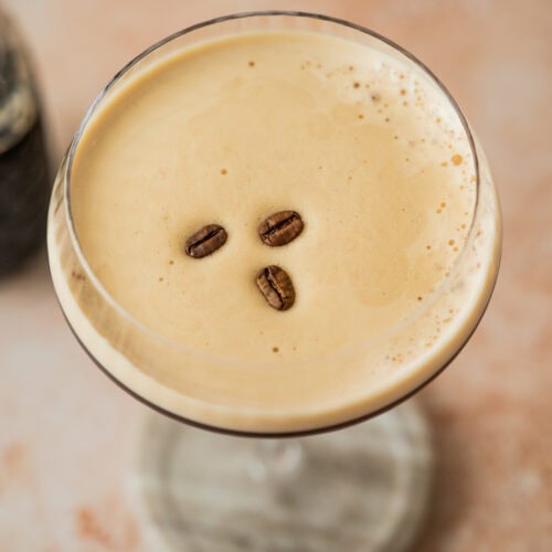 A creamy Virgin Espresso Martini mocktail in a coupe glass, topped with three coffee beans, viewed from above on a light, slightly textured surface.