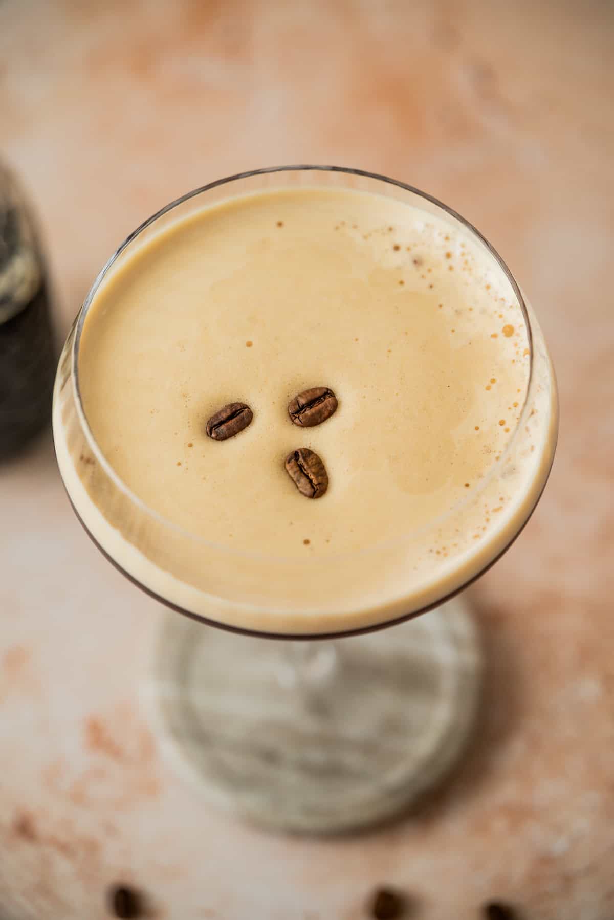 A creamy Virgin Espresso Martini mocktail in a coupe glass, topped with three coffee beans, viewed from above on a light, slightly textured surface.
