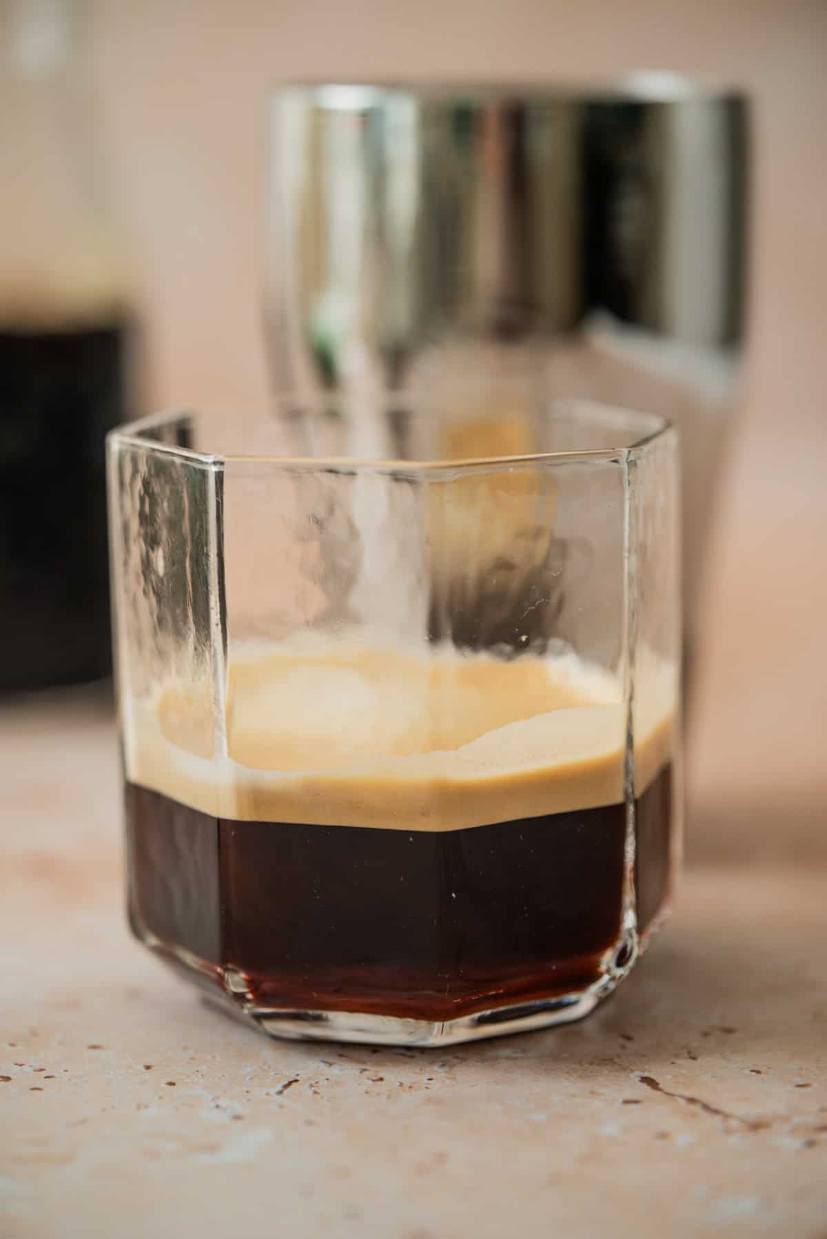 A close-up of a glass with a dark homemade Virgin Coffee Liqueur and a creamy foam layer on top, set on a light surface. A metallic cocktail shaker and a blurred bottle are visible in the background.