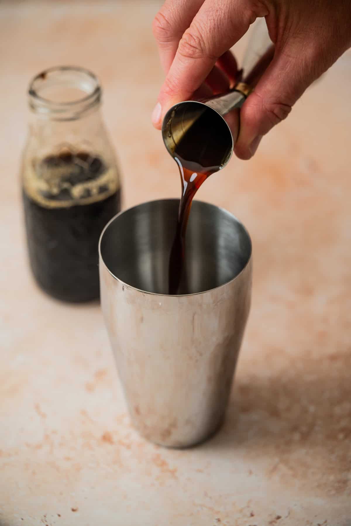 A hand pours a dark liquid from a jigger into a stainless steel cocktail shaker, preparing a Virgin Espresso Martini, with a glass jar of the same dark liquid in the background on a light-colored surface.