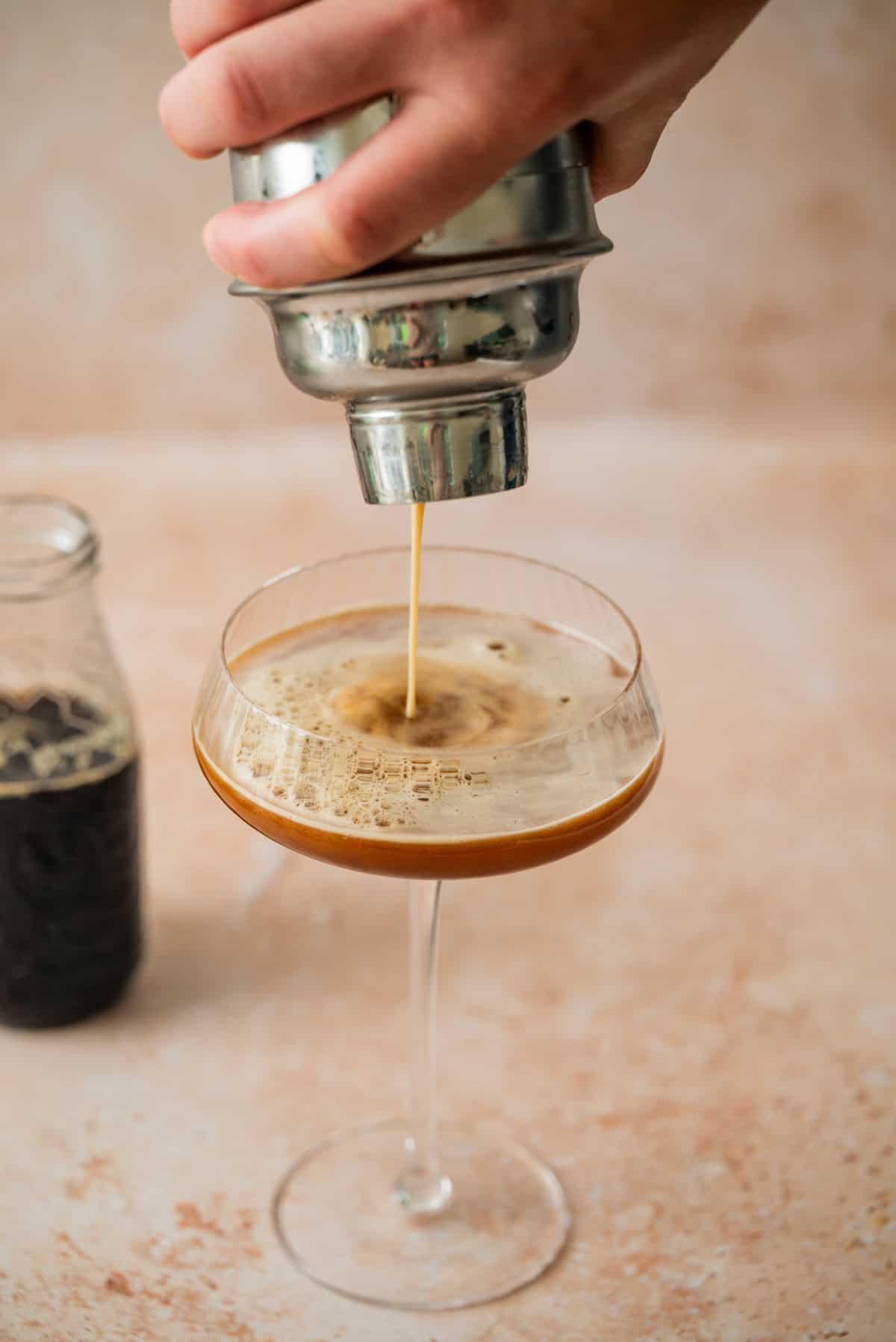 A hand pours a frothy Virgin Espresso Martini from a cocktail shaker into a stemmed glass, with a small bottle of dark liquid in the background.
