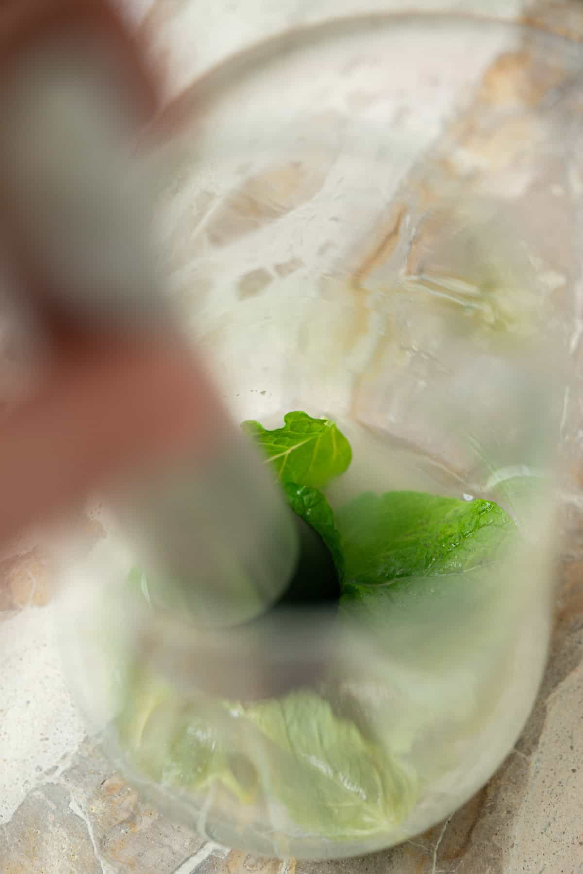 A close-up of fresh mint leaves being muddled in a glass with a muddler on a light-colored countertop.