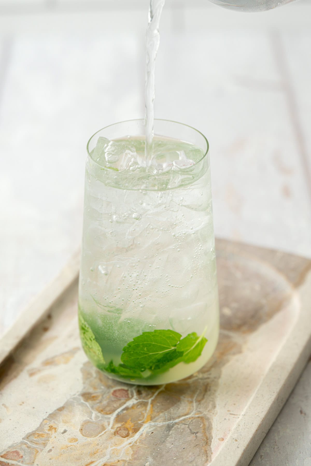 A glass filled with ice, lime juice, and mint leaves is being topped with a clear liquid, possibly sparkling water, on a stone tray with a light background.