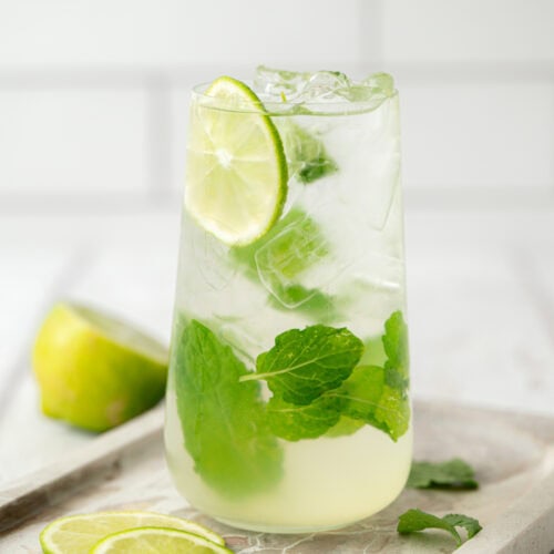 A refreshing glass of mojito with ice cubes, lime slices, and fresh mint leaves, placed on a tray with extra lime slices and mint. A halved lime is in the background. The setting is bright and light.