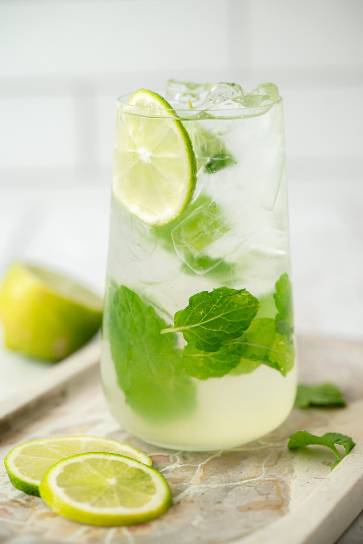 A glass of iced mojito garnished with fresh mint leaves and lime slices, sitting on a marble surface with extra lime slices and mint nearby.