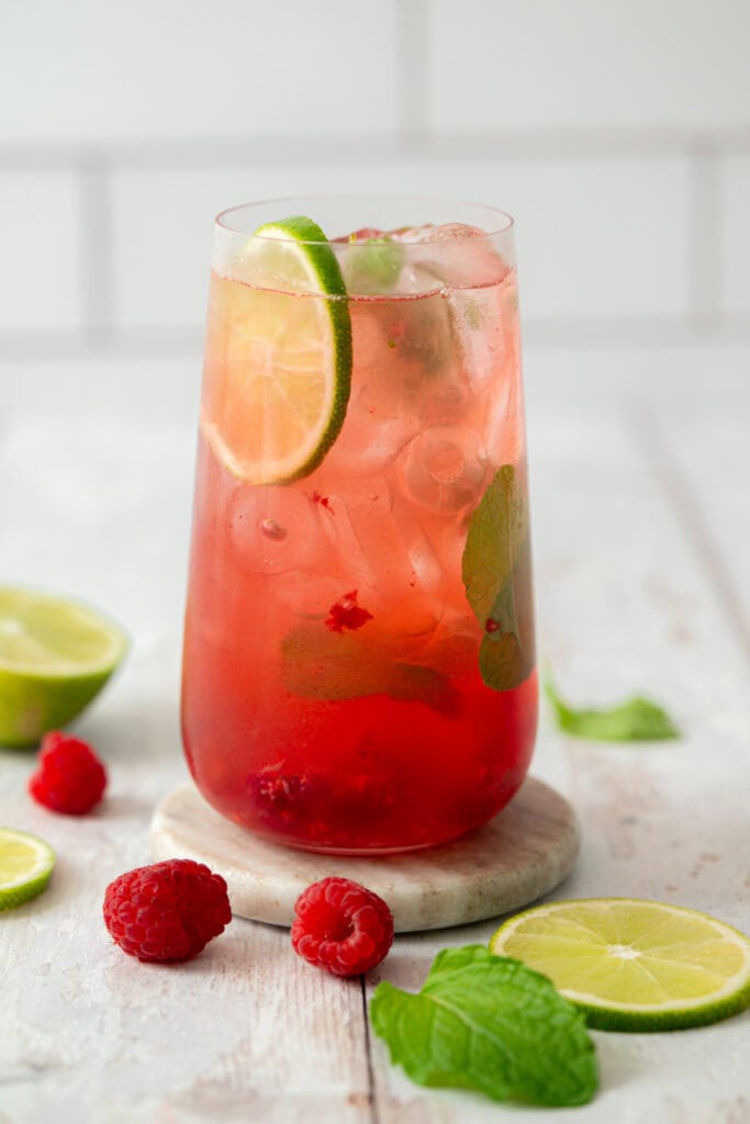 A tall glass of pink raspberry lime drink with ice, garnished with lime slices and mint leaves. Fresh raspberries, lime slices, and mint are scattered on a light surface around the glass.
