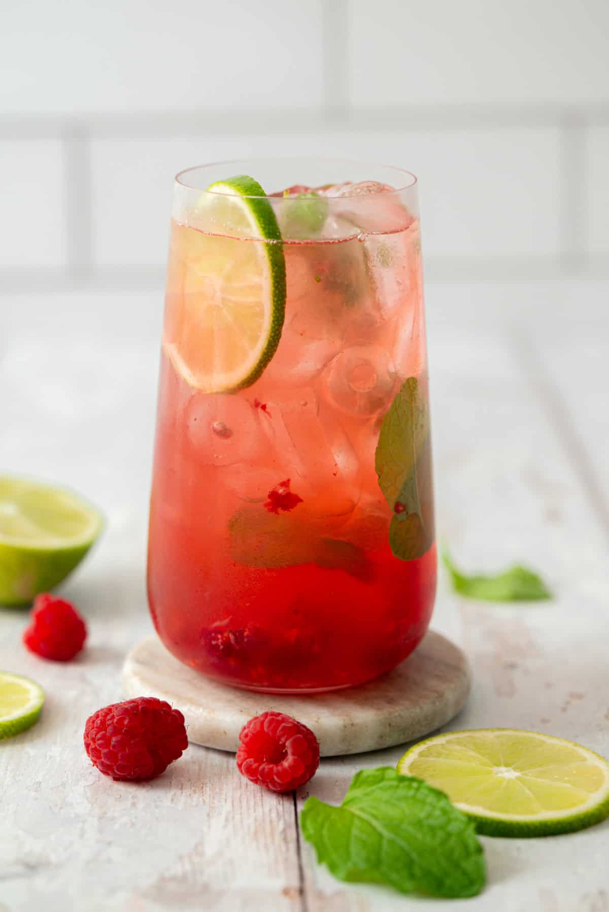A tall glass of pink raspberry lime drink with ice, garnished with lime slices and mint leaves. Fresh raspberries, lime slices, and mint are scattered on a light surface around the glass.