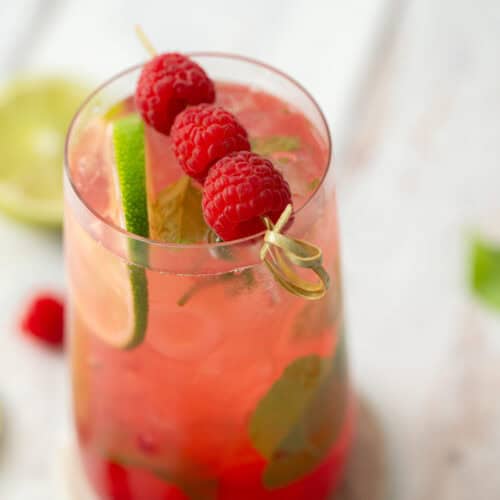 A glass of pink iced drink garnished with fresh raspberries on a skewer, a lime slice, and mint leaves, placed on a white surface with blurred raspberries and lime in the background.