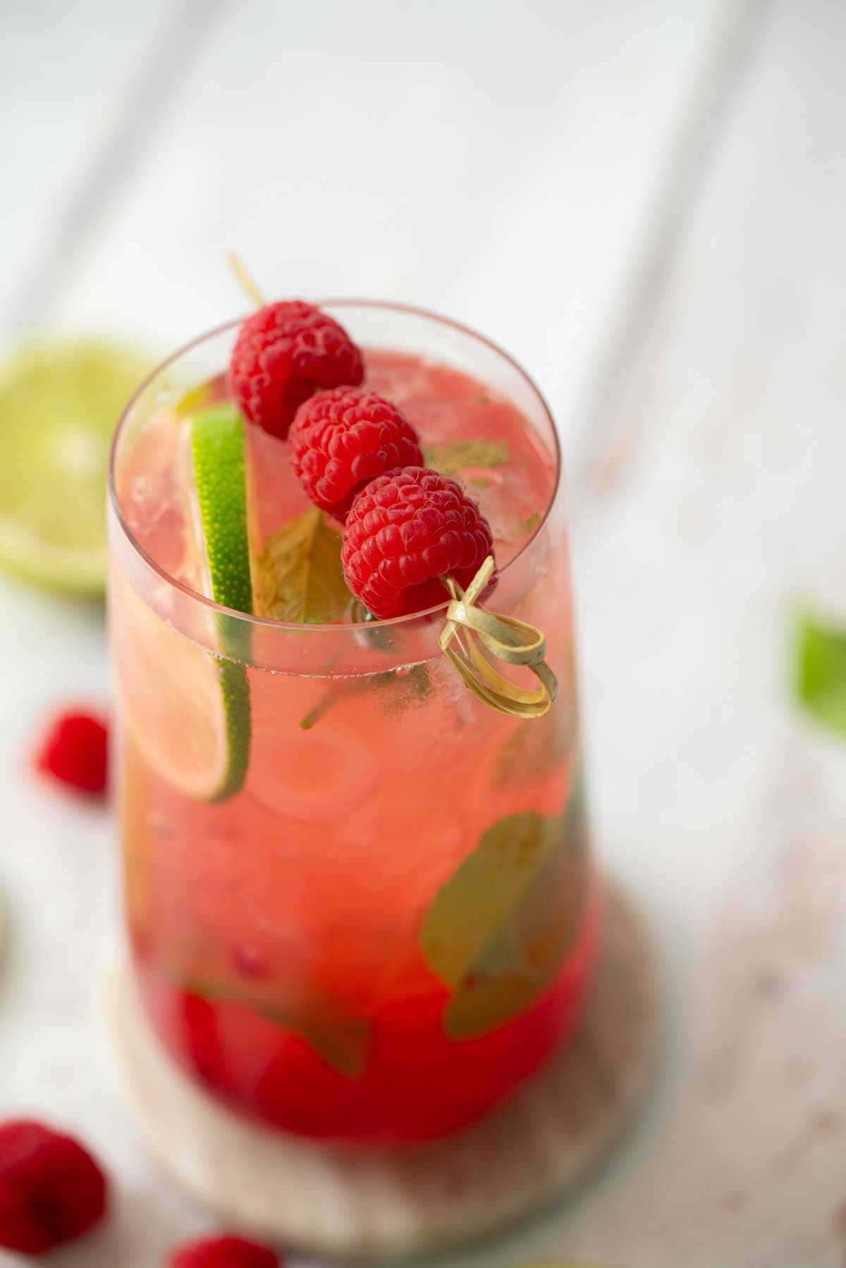 A glass of pink iced drink garnished with fresh raspberries on a skewer, a lime slice, and mint leaves, placed on a white surface with blurred raspberries and lime in the background.