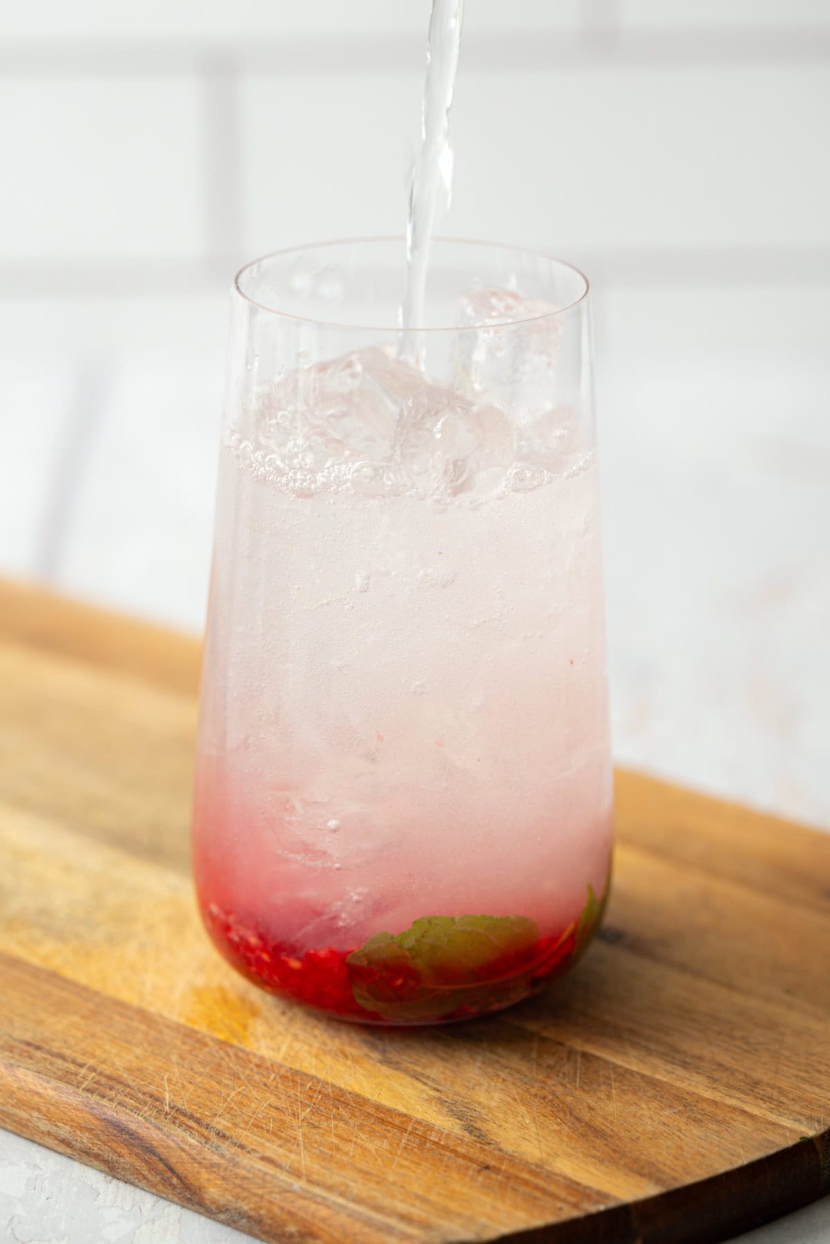 A glass filled with ice, pink liquid, and muddled red fruit and mint at the bottom, being topped off with a clear liquid, sits on a wooden board.