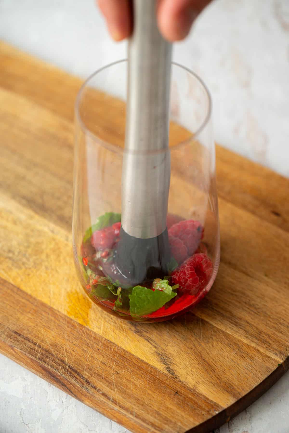 A hand uses a muddler to crush raspberries and mint leaves in a clear glass on a wooden cutting board.