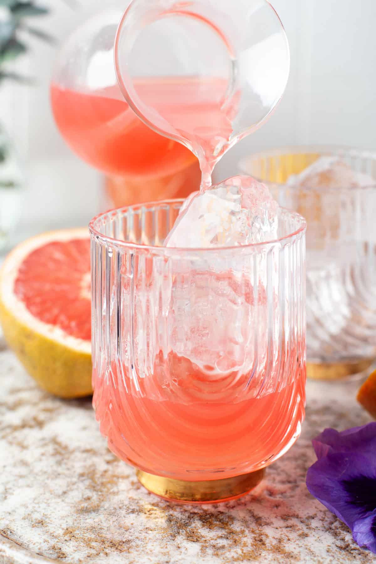 A glass of pink grapefruit drink with ice is being poured from a small pitcher. Half a grapefruit and another glass are in the background, with a purple flower nearby on a light, textured surface.