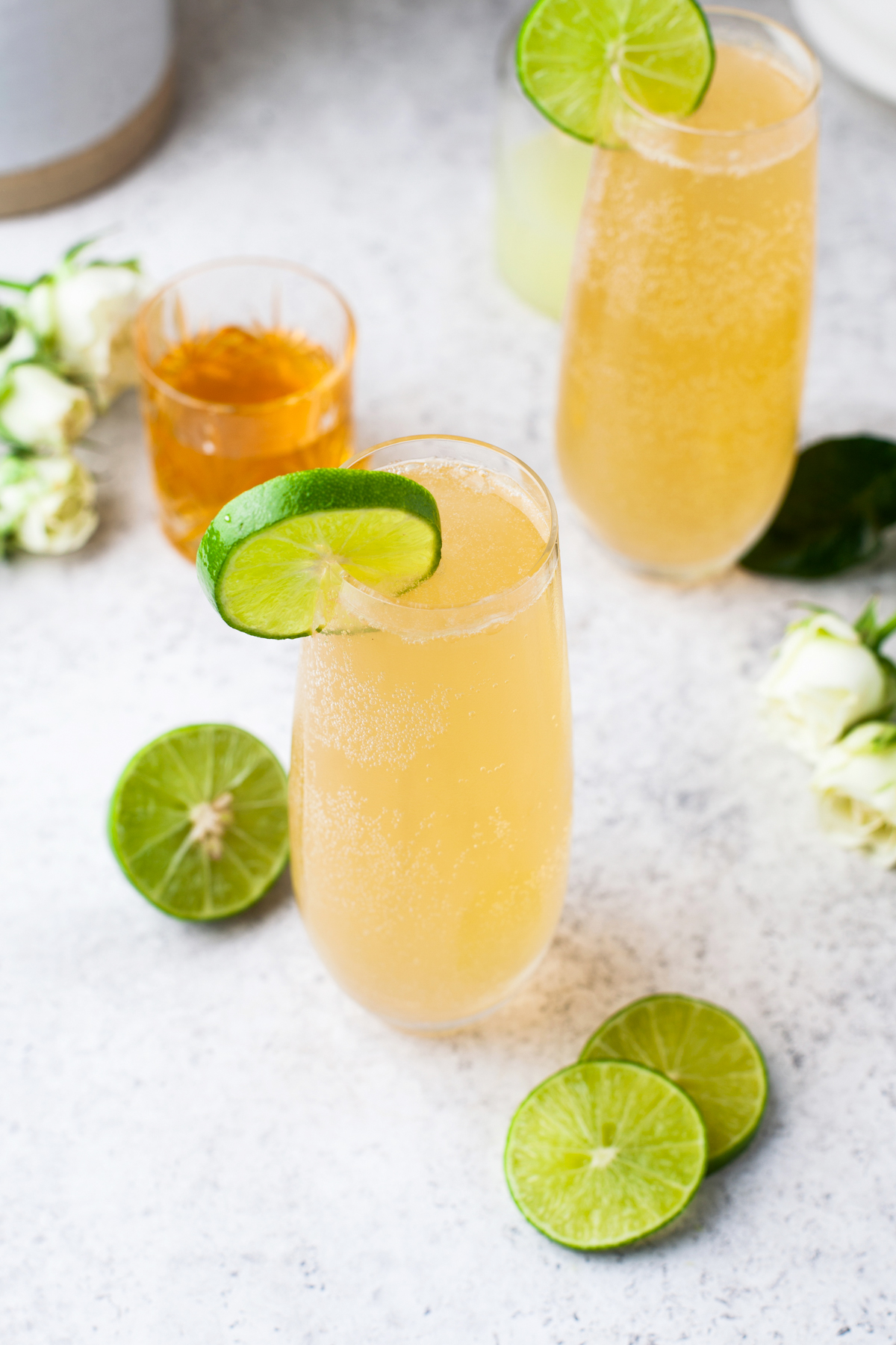 A tall glass of light orange sparkling drink garnished with a lime slice, surrounded by lime halves, lime slices, white flowers, and another glass of the same drink in the background.