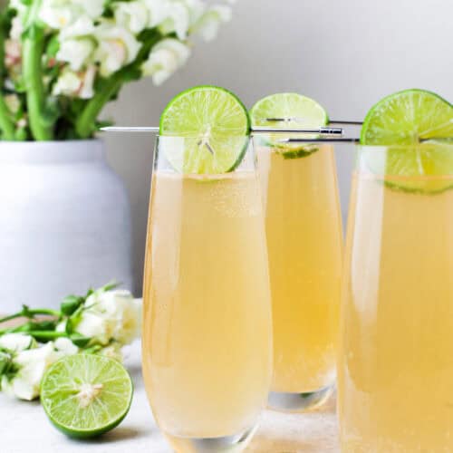 Three tall glasses of a light golden fizzy drink, each garnished with a lime wheel, sit on a white surface with sliced limes and white flowers in the background.