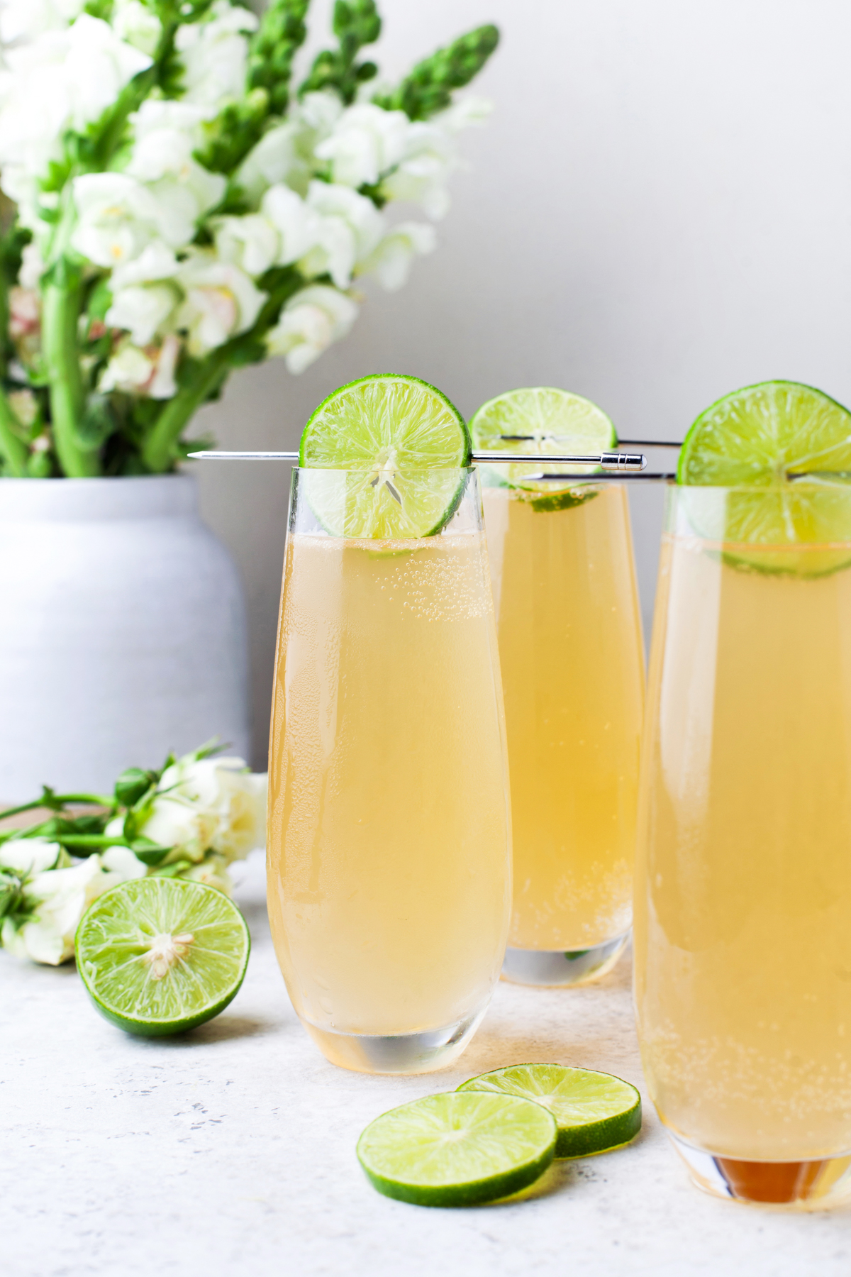 Three tall glasses of a light golden fizzy drink, each garnished with a lime wheel, sit on a white surface with sliced limes and white flowers in the background.