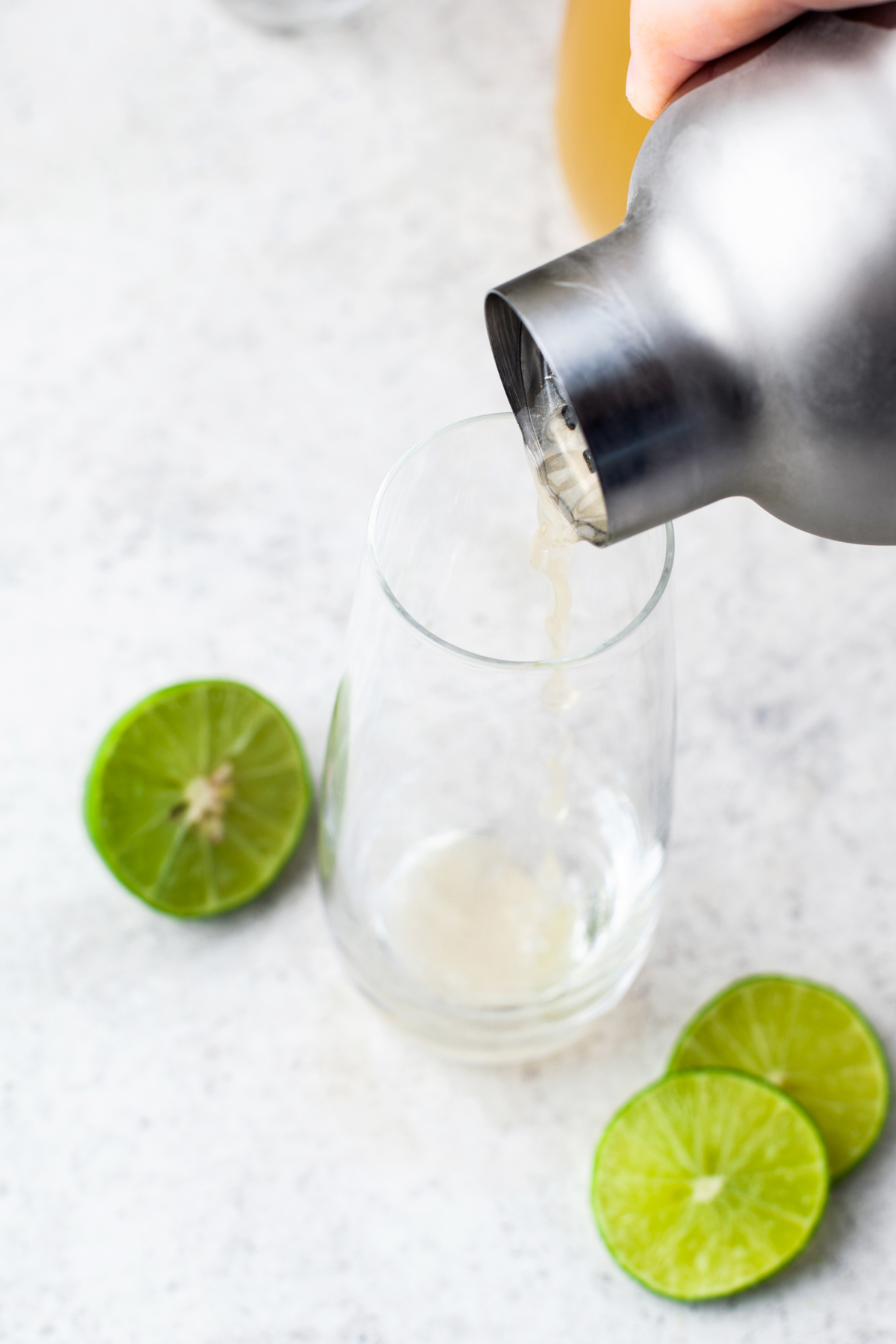 A hand pours liquid from a cocktail shaker into a clear glass, with fresh lime halves and slices on a light-colored surface nearby.