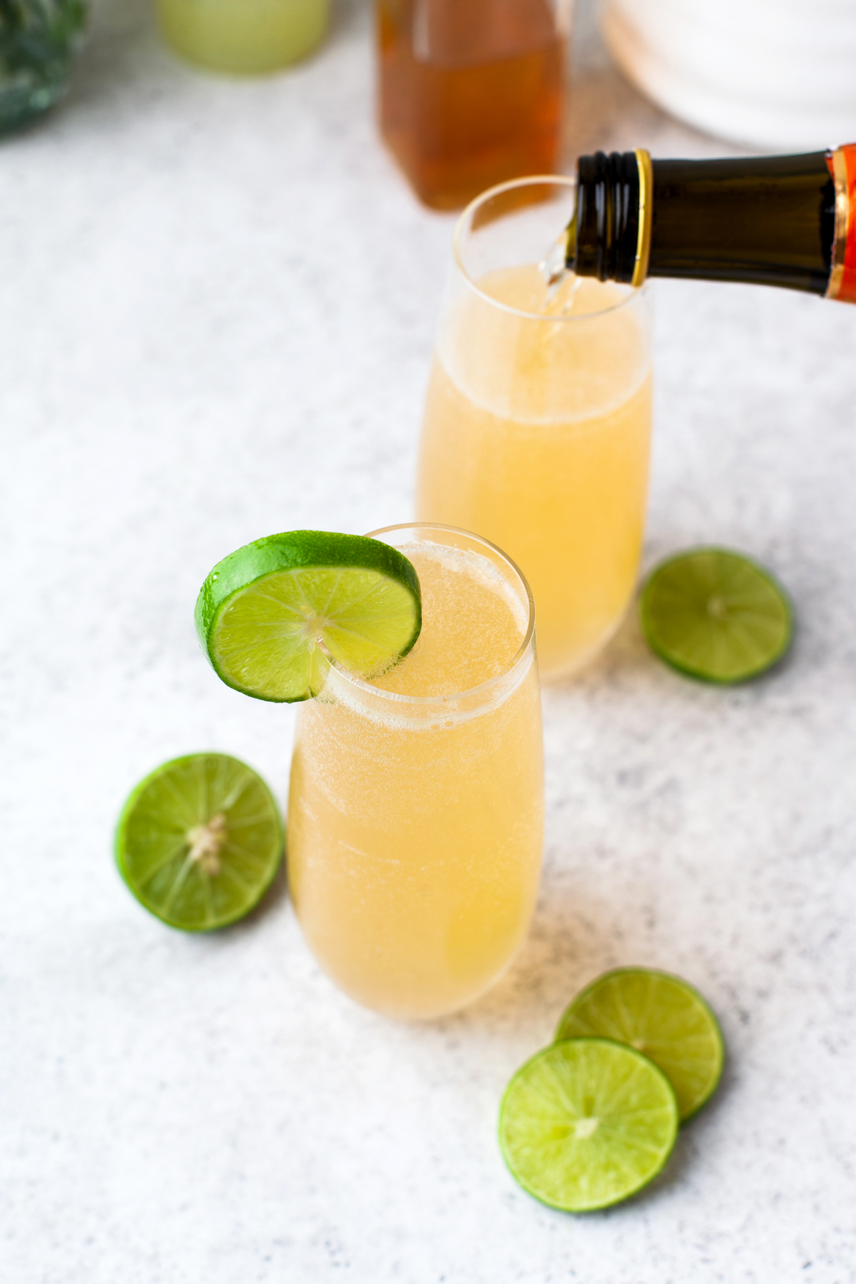Two champagne flutes filled with a the air mail cocktail garnished with lime slices. One glass is being topped with sparkling wine. More lime slices are scattered on the light-colored surface around the glasses.