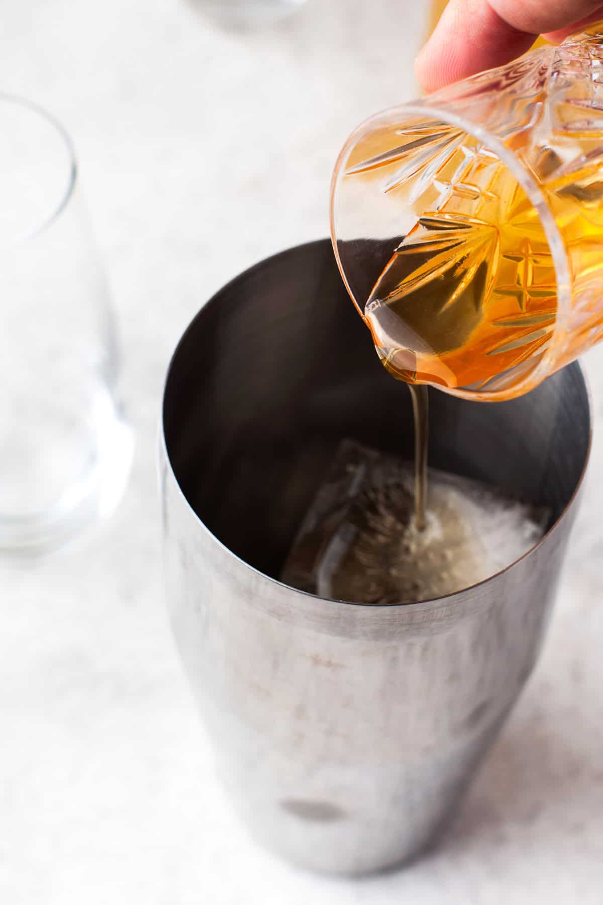 A hand pours a golden-colored liquid from a patterned glass into a metal cocktail shaker containing ice cubes, with an empty glass nearby on a light surface.