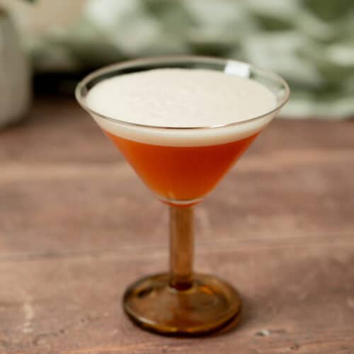A brown cocktail with a frothy, creamy foam on top is served in a vintage-style martini glass, set on a wooden surface with a blurred green patterned background.