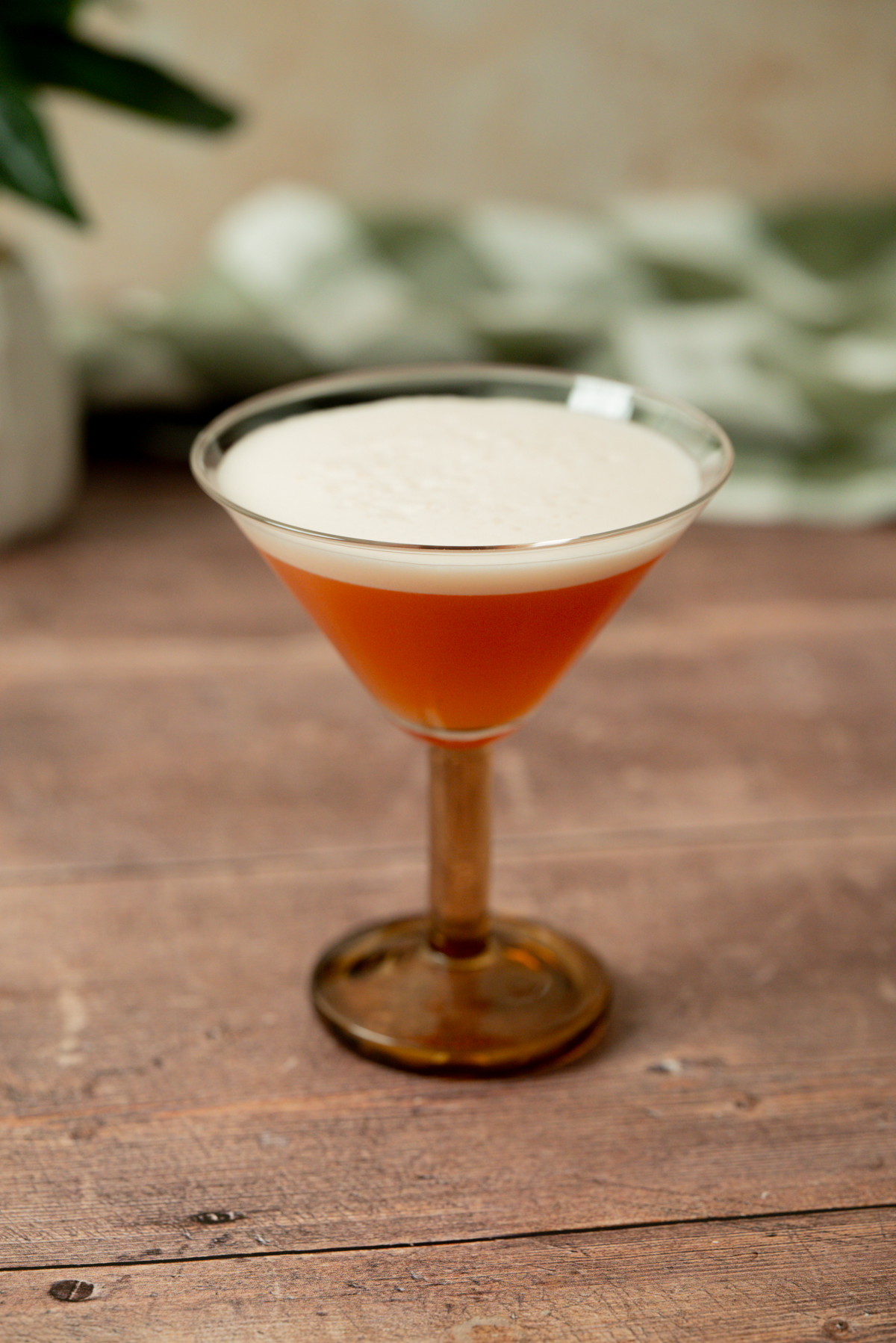 A brown cocktail with a frothy, creamy foam on top is served in a vintage-style martini glass, set on a wooden surface with a blurred green patterned background.