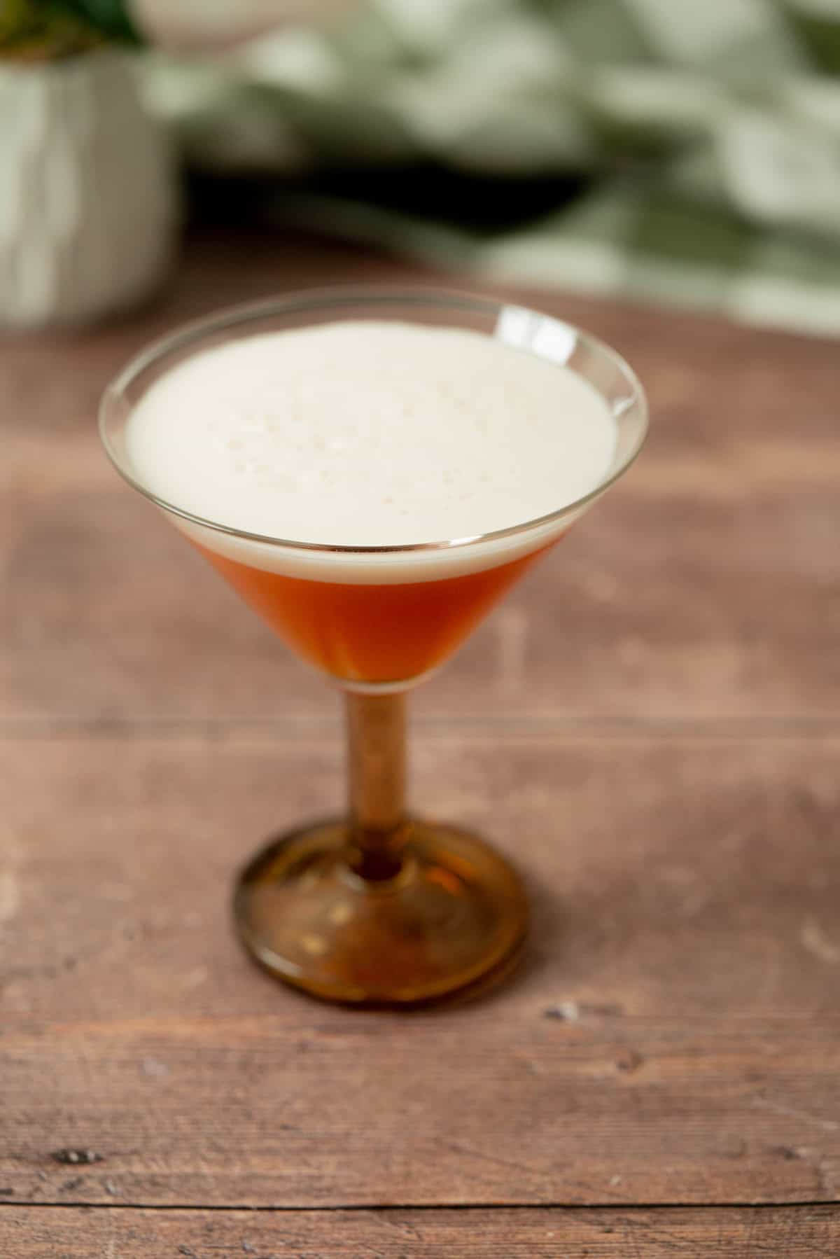 A cocktail in a brown-stemmed martini glass sits on a wooden surface, topped with a layer of foam. The background is softly blurred with hints of green and white.