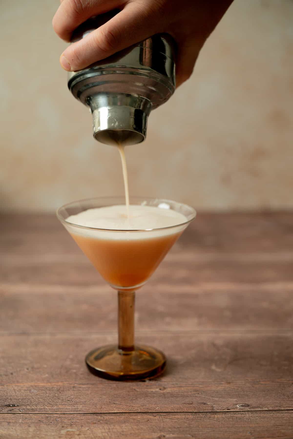 A hand pours a frothy cocktail from a metal shaker into a martini glass on a wooden surface, with a softly blurred beige background.
