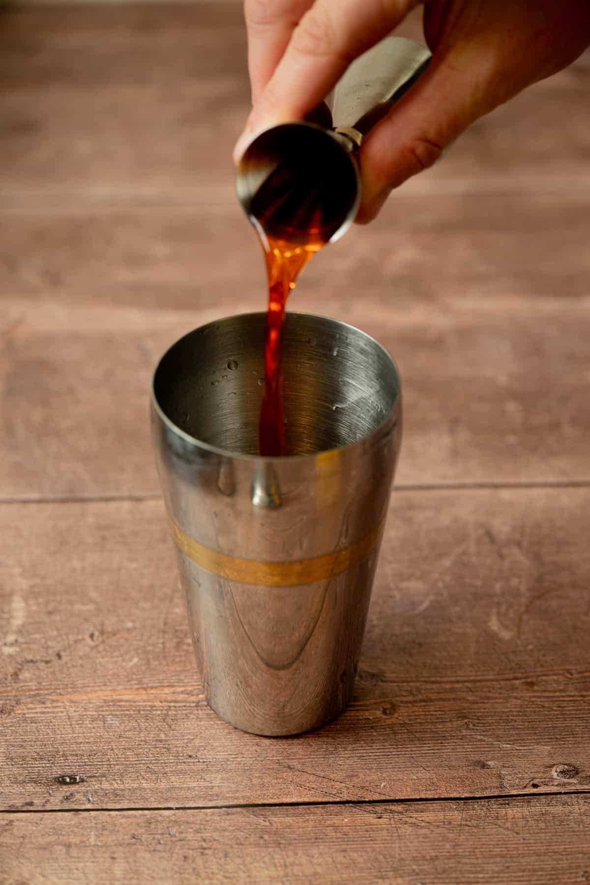 A hand pours a dark liquid from a small metal jigger into a stainless steel cocktail shaker on a wooden surface.