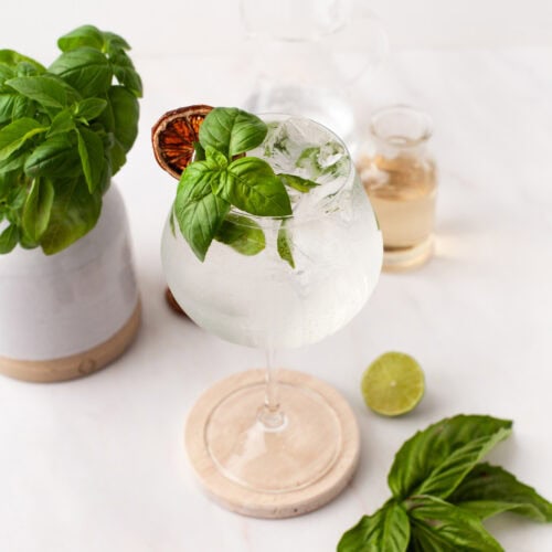 A glass of clear cocktail with ice, garnished with fresh basil leaves and a dried citrus slice, sits on a round coaster. Surrounding the glass are fresh basil, a halved lime, and small bottles on a white surface.