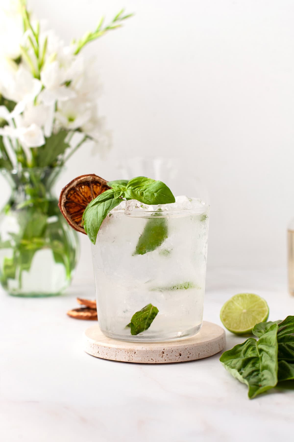 A glass of clear cocktail with ice, basil leaves, a dried lime slice garnish, and fresh lime and basil on the side. A vase of white flowers is blurred in the background.