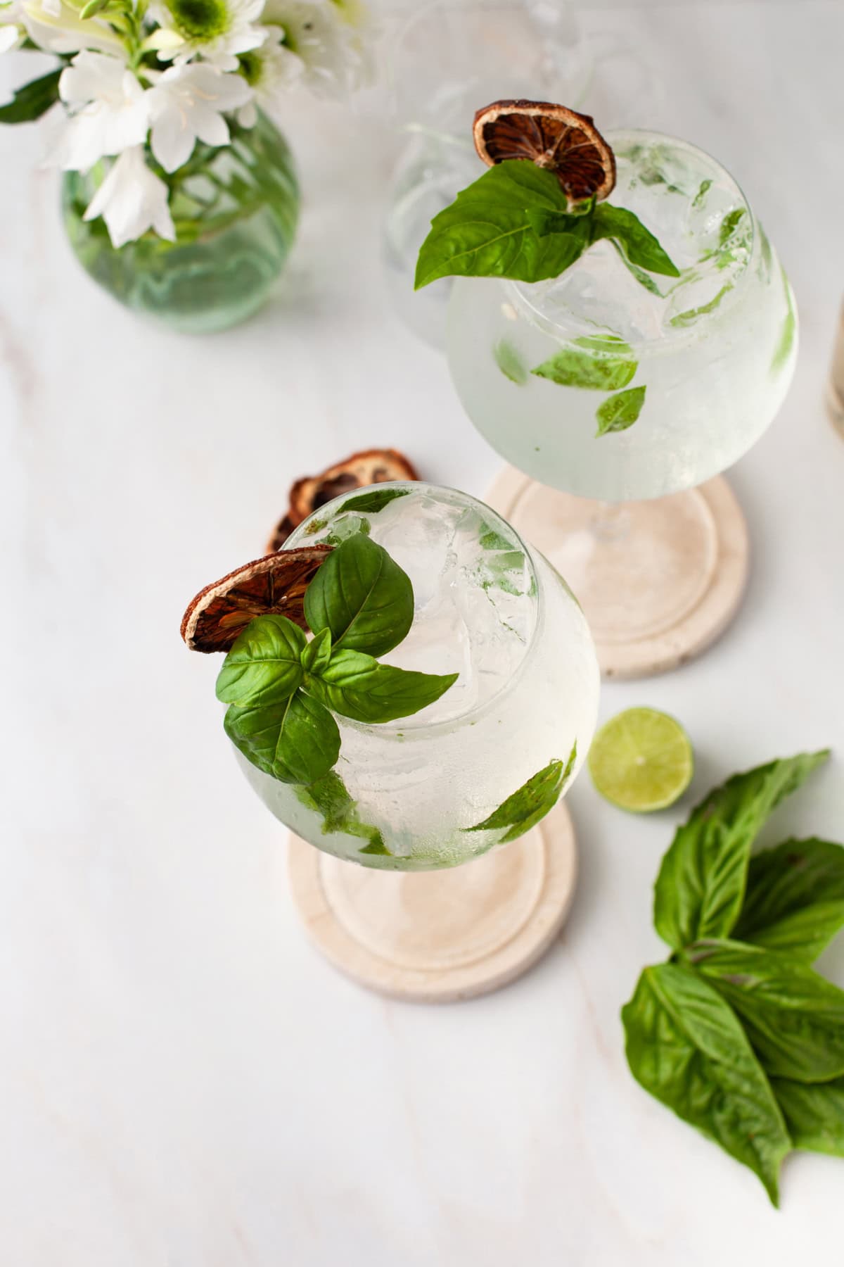 Two glasses of a clear cocktail with ice, fresh basil leaves, and dried citrus garnish on round coasters. A sliced lime and basil leaves are nearby, with a vase of white flowers in the background on a light surface.