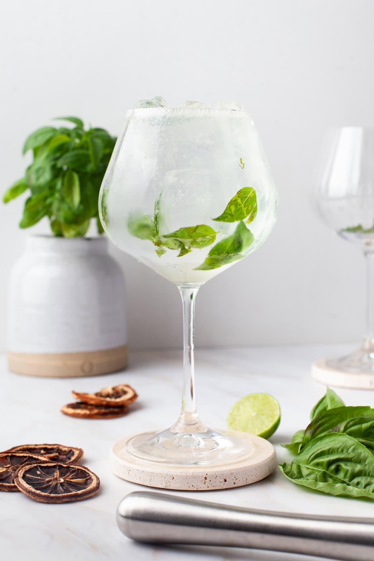 A gin and tonic in a large, round glass garnished with fresh basil leaves and ice, sits on a coaster. In the background are a basil plant, dried citrus slices, and half a lime on a white surface.