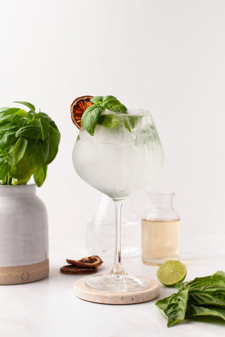 A gin and tonic garnished with fresh basil leaves and a dried citrus slice sits on a coaster. Nearby are a basil plant, a small glass bottle with clear liquid, a slice of lime, and loose basil leaves.