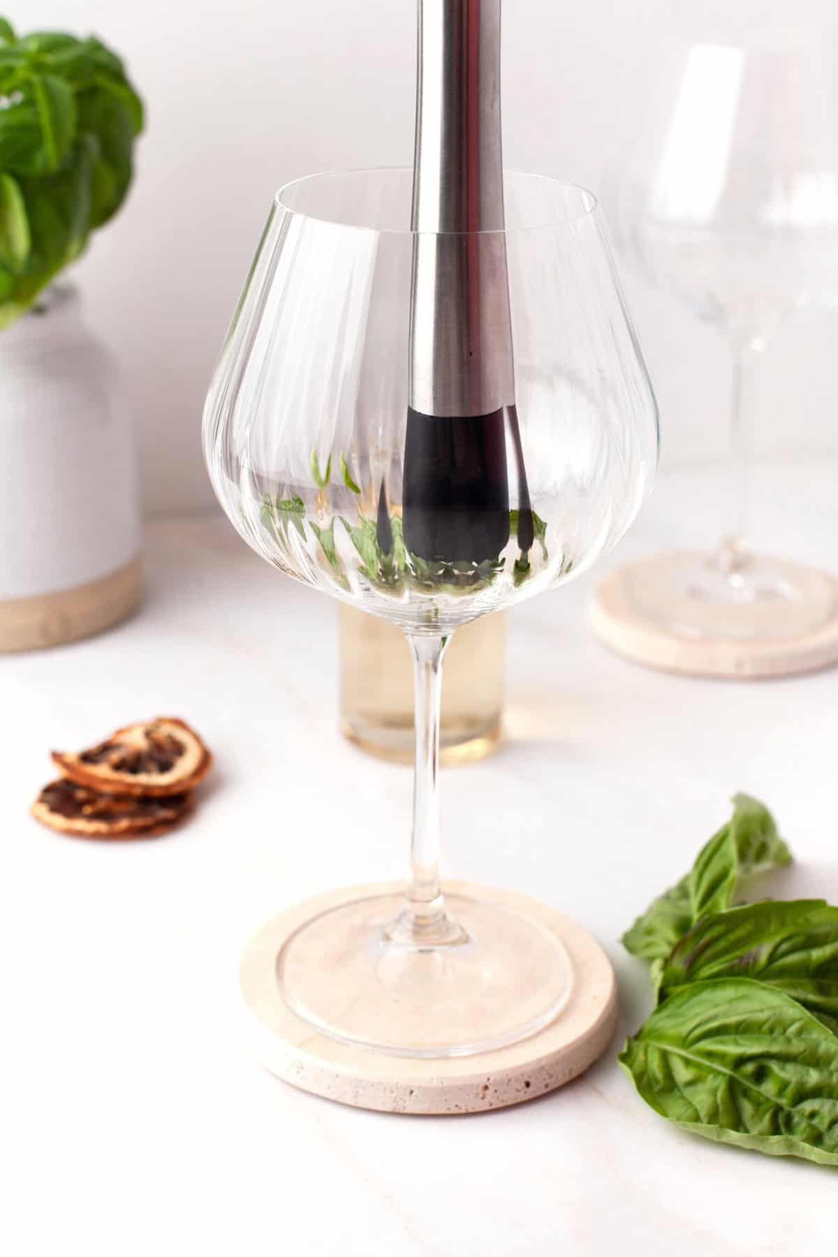 A muddler pressing fresh basil leaves in a large wine glass on a coaster, with sprigs of basil and dried citrus slices nearby on a white surface.